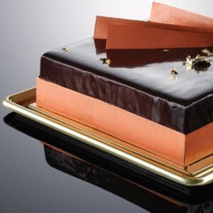 A rectangular cake with a glossy chocolate glaze is elegantly presented on a golden tray. The sides feature an orange layer and gold leaf accents, reminiscent of artisanal gelato craftsmanship. Three thin chocolate pieces are artfully laid diagonally on top, reflected in the tray's shiny surface.