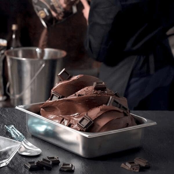 Close-up of a stainless steel container filled with rich, creamy chocolate gelato, garnished with squares of dark chocolate. In the background, a person wearing dark clothing pours melted chocolate from a pot into a bucket in an industrial kitchen setting as if they were checking My Account for perfection.