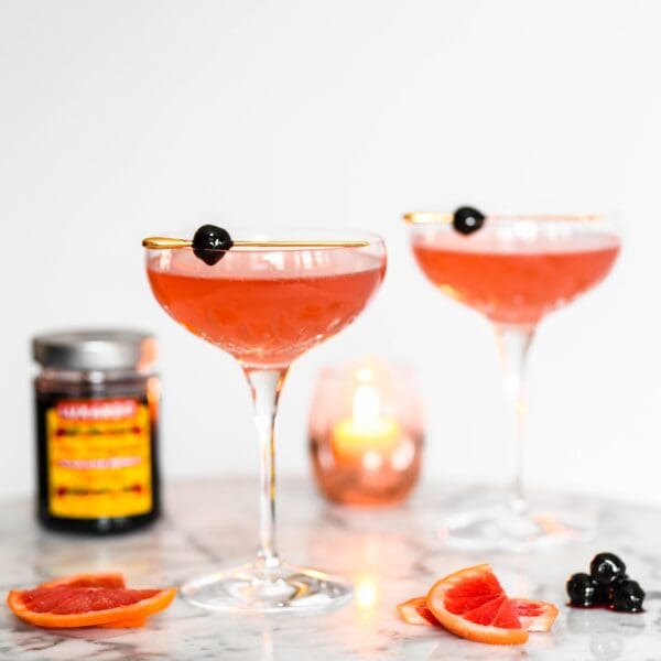 Two coupe glasses with pink cocktails are garnished with Luxardo Gourmet Maraschino Cherries in Syrup on gold rims. A lit candle, sliced blood oranges, and a jar of syrup-infused cherries sit on the marble surface.