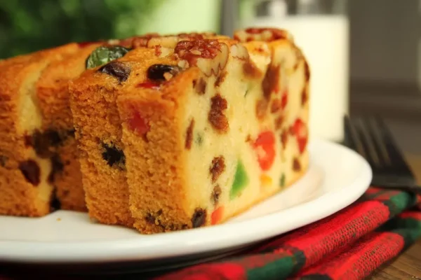 Close-up of a plate featuring two slices of fruitcake adorned with preserved candied fruit, such as Ambrosio candied pears. The golden-brown crust of the cake provides a stunning contrast. In the background, a fork and a glass of milk rest on a plaid cloth.