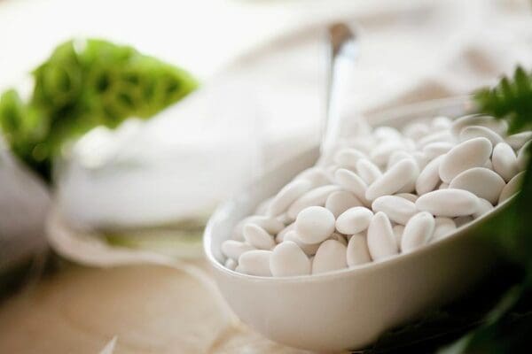 A white bowl filled with Ambrosio Confetti Sugar Covered Almonds (12x1Kg case) sits on a table. In the background, blurred green leaves enhance the inviting ambiance, while a silver spoon rests in the bowl, all bathed in soft, warm light.