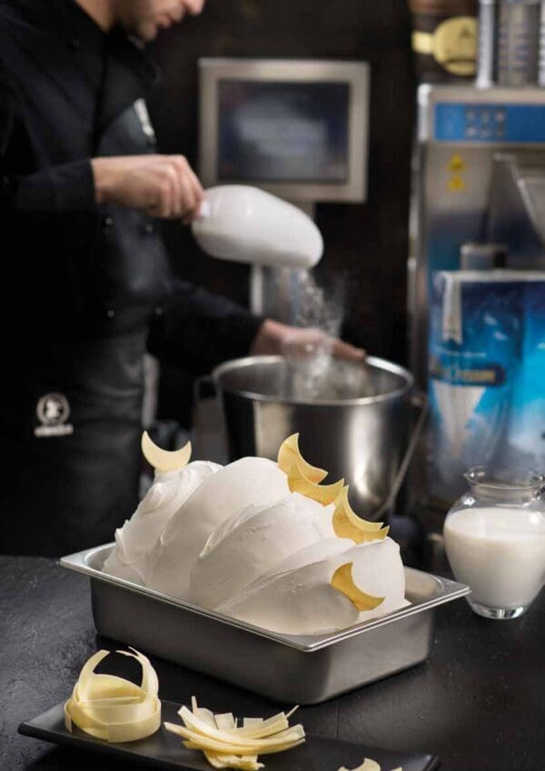 A person in a black uniform is pouring milk into a large metal mixing bowl, crafting the perfect ice cream base using Elenka Ice Cream Base Ingredients Novacrem 50. In the foreground, a container of smooth, white gelato garnished with decorative white chocolate curls is placed on a dark surface. A jug of milk and a tray with more white chocolate curls are nearby.