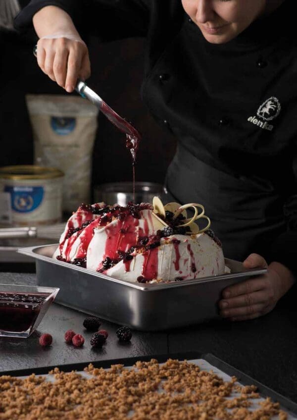 A pastry chef decorates a white, oval-shaped cake with dark red sauce and blackberries. The cake sits in a metal tray. Nearby, there are scattered berries, a glass container with more sauce, and a tray of crumbled topping next to an Elenka Ice Cream Base Ingredients Novacrem 50 (20x1Kg case) ready for the final touch.