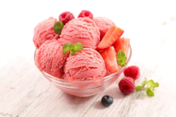 A glass bowl filled with scoops of Elenka Strawberry Sorbet Gelato Paste is garnished with fresh mint leaves, raspberries, strawberry slices, and a blueberry. The bowl is placed on a white wooden surface, adding a rustic charm to the vibrant dessert.