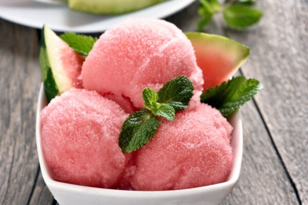 A white bowl holds three scoops of vibrant pink watermelon sorbet, decorated with fresh mint leaves and small watermelon slices. Positioned on a wooden table, the bowl is further accentuated by a green leafy garnish. This refreshing sorbet is effortlessly prepared using Elenka Instant Watermelon Base for Granitas, Sorbets and Ice Cream (10x1.5Kg Case).