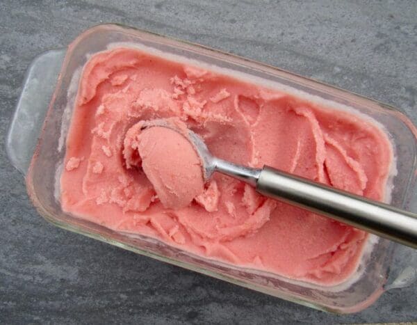 A glass container filled with pink-colored sorbet, crafted from Elenka Instant Watermelon Base for Granitas, Sorbets and Ice Cream. A metal ice cream scoop holds a portion of the delectable sorbet and rests on top of the remaining treat. The serene grey background complements the 10x1.5Kg case of sweet indulgence perfectly.