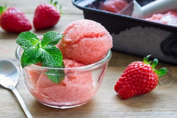 A bowl of Elenka Strawberry Sorbet Gelato, garnished with a sprig of fresh mint, accompanied by a spoon, a whole strawberry, and a tray of Elenka Strawberry Sorbet Gelato Paste (4x3Kg case) in the background. The sorbet has a vibrant, pinkish-red color and appears refreshing.