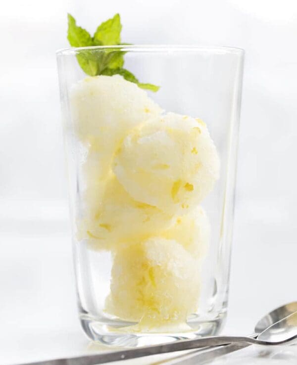 A clear glass filled with three scoops of gelato made from Elenka Lemon 50 Powder Base (20x1Kg case), garnished with a fresh mint leaf. The glass is set against a soft white background, while a spoon is visible in the foreground, inviting you to indulge in this tangy delight.