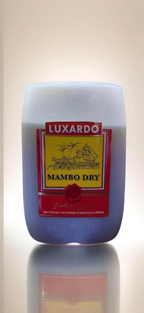 A jug of Luxardo Baking Liquor Rum 'Mambo Scuro' with a light gray cap. The label depicts a ship and palm trees in yellow and red colors. The text on the label is in Italian with "Luxardo" prominently displayed in large red letters at the top, evoking a vintage baking ingredients packaging style. The bottle stands illuminated against a bright background.