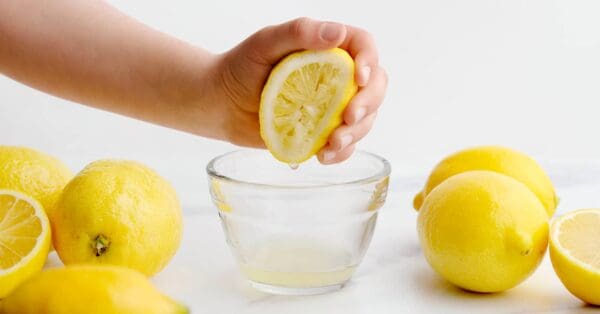 A hand squeezes half a lemon over a small glass bowl of Elenka Agrolina Ciaculli Lemon Juice Concentrate for Sorbet, enhancing its vibrant essence. Whole lemons rest on a white surface, setting the perfect stage to prepare this sorbet concentrate.