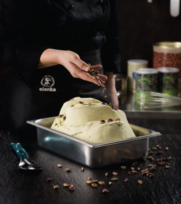 A person in a black apron is sprinkling pistachios over a large container of pistachio gelato. The gelato is in a stainless steel container on a dark countertop, with additional containers and utensils in the background.