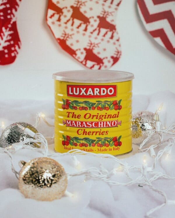 A can of Luxardo Gourmet Maraschino Cherries in Syrup (4x5.6Kg case) sits on a snowy surface decorated with string lights and glittery ornaments, while festive stockings hang in the background, creating a charming holiday scene.