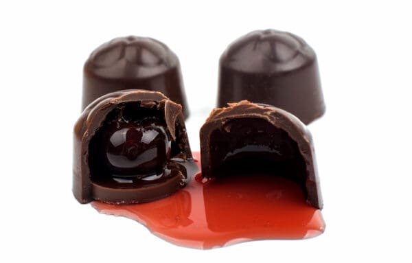 Four dark chocolate bonbons are arranged on a white background, with one cut in half to reveal the enticing filling of Luxardo Gourmet Maraschino Cherries in Syrup, with the syrup gently oozing out.