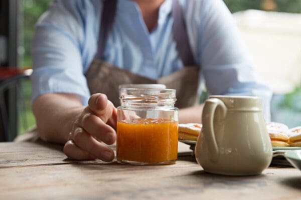 A person in a light blue shirt and apron reaches for a jar of orange marmalade on a wooden table that also holds Lazzaris Mostarda Preserved Fruits with Mustard (2x5Kg case). Nearby, a white creamer and a plate with pastries invite indulgence, while the softly blurred window view adds tranquility.