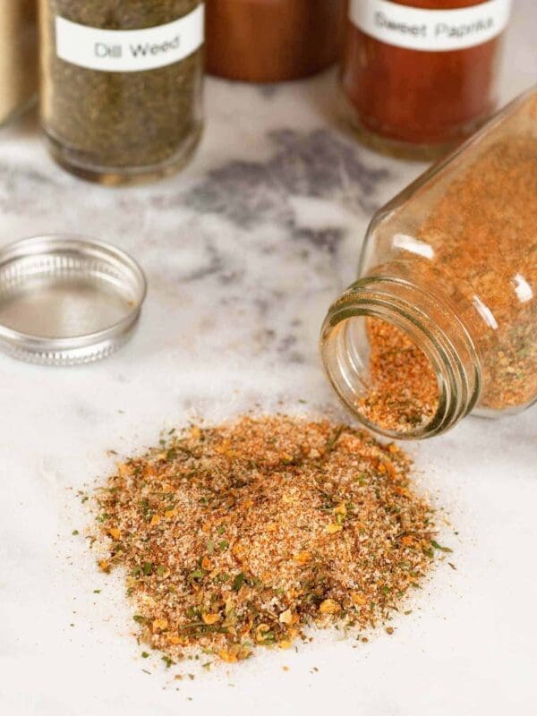 A jar of spice mix is tipped over on a marble surface, spilling its contents like a grocery store mishap. Surrounding the jar are other spice containers, including ones labeled "Dill Weed" and "Sweet Paprika.