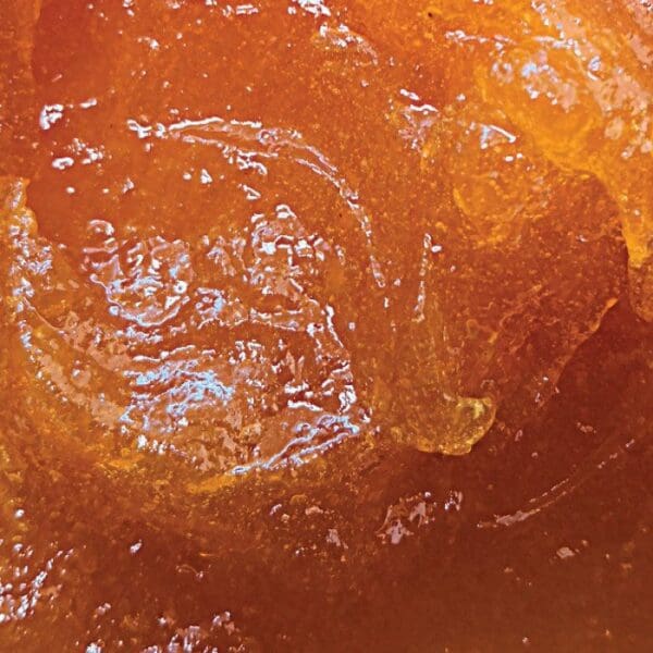 Close-up view of a thick, amber-colored substance with a glossy, slightly uneven surface. The texture has a gel-like appearance, creating light reflections and subtle variations in color ranging from deep orange to golden—reminiscent of the luscious filling found in an Elenka Apricot Jam Preserve for Crostata, Croissants, Pastries and Baked Goods (1x12Kg).