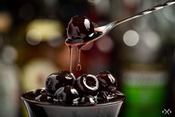 A spoon holds a glossy cherry dripping syrup above a bowl filled with Luxardo Gourmet Maraschino Cherries in Syrup, set against a softly blurred backdrop of various out-of-focus bottles.