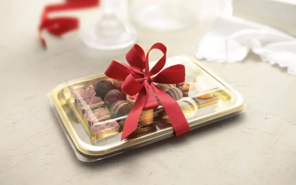 A decorative Alcas Kadò Tray, filled with assorted chocolates and sweets and tied with a bright red ribbon, sits on a light-colored surface. In the background, a soft white cloth and red ribbon are softly blurred.