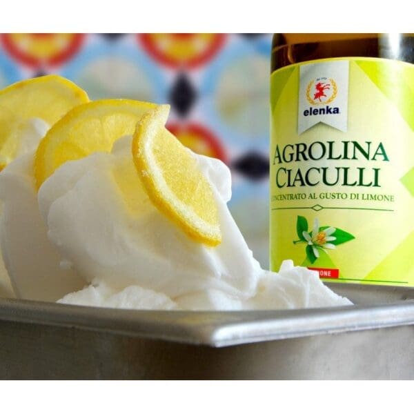 A metal container filled with fluffy white gelato, garnished with lemon slices, is set against a colorful backdrop. Behind it stands a bottle of Elenka Agrolina Lemon Gelato Concentrate Flavors for Sorbet (6x2.75Kg case) featuring a vibrant lemon design.