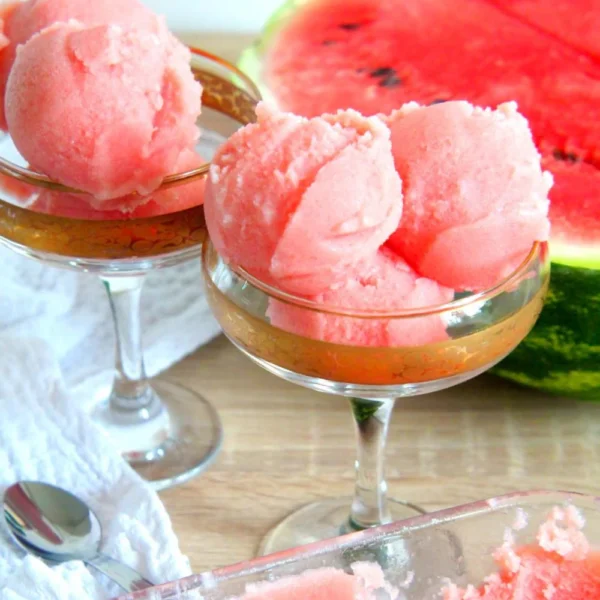 Two glass dishes hold scoops of pink watermelon sorbet made with the Elenka Gelato Watermelon Instant Base for Sorbets, while a halved watermelon sits in the background on a wooden surface, accompanied by a spoon and white cloth.