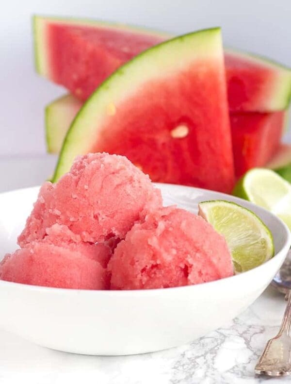 A white bowl contains three scoops of refreshing watermelon sorbet made with Elenka Gelato Watermelon Instant Base for Sorbets, garnished with a lime wedge. Fresh watermelon slices are placed on a marble surface behind the bowl.