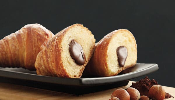 Two croissant cones filled with Elenka Farcitella Chocolate Cream Filling are elegantly presented on a black tray. Whole hazelnuts and cocoa powder scattered in the foreground on wood enhance their appeal against a dark backdrop, reminiscent of indulgent cakes.