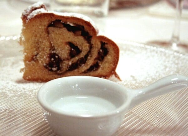 A piece of Vogrig Gubana Cake from Friuli-Venezia (6x850gr case) rests stylishly on a clear glass plate, sprinkled with powdered sugar, and paired with a small white bowl filled with sauce.