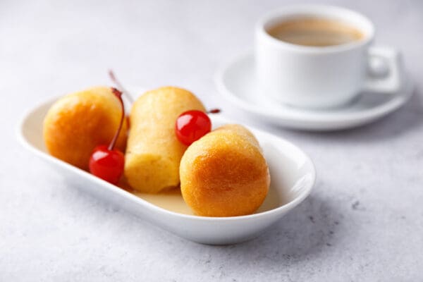 Three petite cakes, each garnished with cherries, are artfully displayed on a white plate, crafted using Luxardo Flavors' 'Mambo Scuro' rum liquor soaks for sponge and cream cakes. A cup of coffee adds to the scene, resting on a light gray surface in the background.