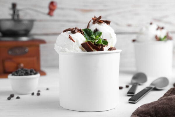 A white paper cup filled with vanilla ice cream made from Elenka Ice Cream Base Ingredients, garnished with chocolate shavings and a sprig of mint on a light wooden table. In the background, there are another cup with ice cream, two spoons, and a small bowl with chocolate chips.