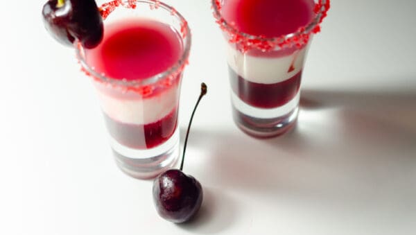 Two layered red and white cocktails in shot glasses, rimmed with red sugar and garnished with Luxardo Gourmet Maraschino Cherries in Syrup. One Luxardo cherry rests on the table beside the glasses, enhancing the vibrant and festive display.