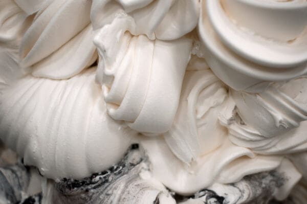 Close-up image of swirling, creamy white texture that appears to be soft-serve ice cream or whipped cream, crafted with Elenka Ice Cream Base Ingredients. Streaks of darker color near the bottom suggest a mix of flavors. The texture is smooth and fluffy with intricate folds and swirls.