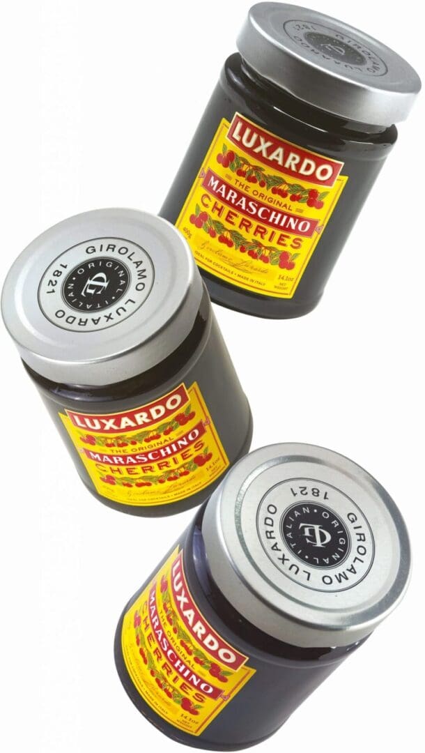 Three jars from a Luxardo Maraschino Cherries 12x400ml case are shown at various angles, featuring black lids and vibrant yellow labels with red and white text. The detailed labels prominently display the Luxardo brand, emphasizing its rich heritage.