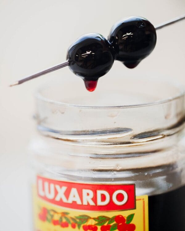 Two dark cherries on a skewer drip juice over an open jar from a "Luxardo Maraschino Cherries (12x400ml case)," highlighting the glossy cherries and jar's edge with a blurred background.