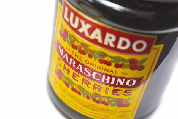 Close-up of a Luxardo Maraschino Cherries 400ml jar label featuring vibrant red cherries and the phrase "The Original Maraschino Cherries." Cherry illustrations embellish the label, with a yellow-to-red gradient accentuating the prominent "Luxardo" at the top.