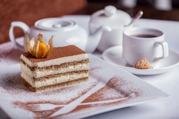 A tiramisu slice, subtly infused with Luxardo Rum Mambo Dry Aroma, sits elegantly on a white plate with cocoa powder art shaped like cutlery. Nearby, a teapot accompanies a teacup on a saucer holding a sugar cube, enhancing the charm of this delightful dessert setting.