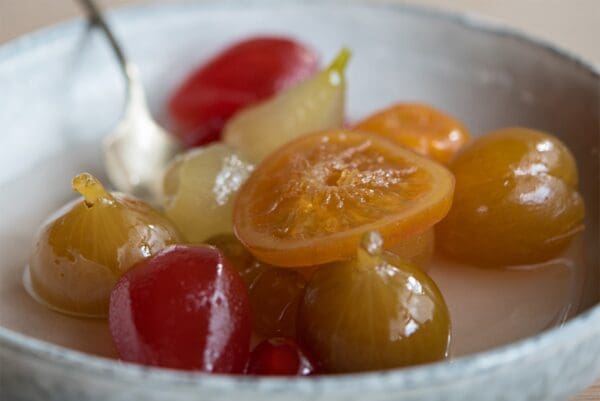 A bowl brimming with Lazzaris Mostarda Preserved Fruits with Mustard (2x5Kg case) displays an array of vibrant candied fruits, featuring a sliced orange segment and red and green cherries, all shimmering in syrup. A spoon sits invitingly within the bowl.