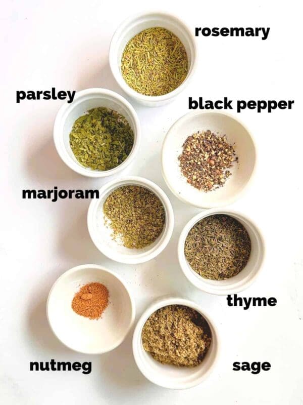 Seven small bowls from a grocery display are filled with various dried herbs and spices, arranged on a white background. Each bowl is labeled: rosemary, parsley, marjoram, nutmeg, black pepper, thyme, and sage.