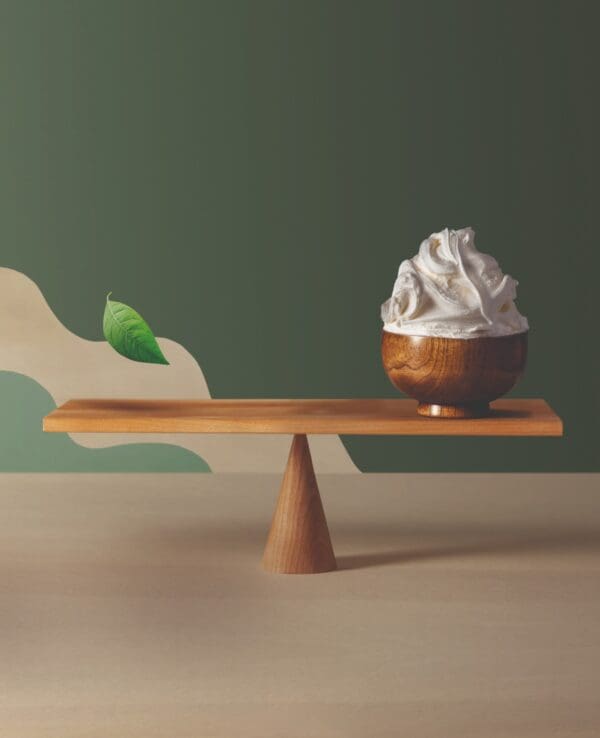 A wooden pedestal balancing a small wooden bowl filled with whipped cream. The background is a two-tone wall, green and cream, with a decorative leaf element on the green side. The pedestal has a modern, minimalist design, reminiscent of Italian products known for their elegance and craftsmanship.