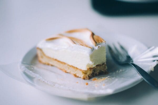 A partially eaten slice of lemon meringue pie sits on a white plate with a fork next to it. The pie has a golden-brown crust, a creamy lemon filling, and a fluffy, lightly browned meringue topping. The flavors are enhanced by delicate extracts, adding depth. The background is blurred and predominantly white.