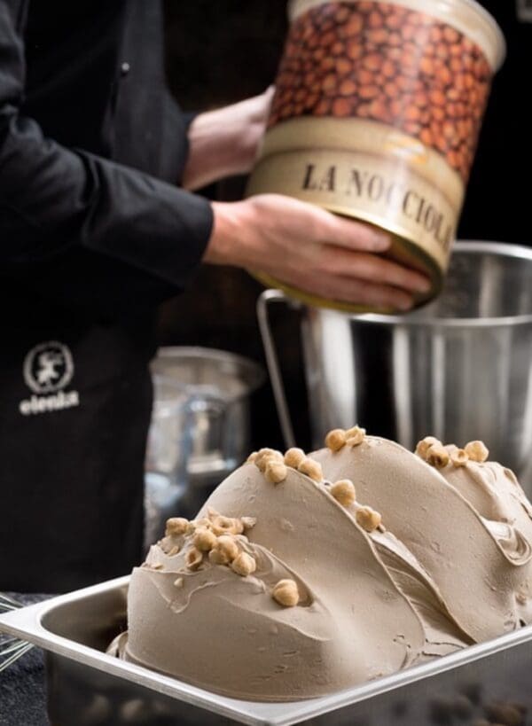 A person in a black apron prepares hazelnut gelato. In the foreground, a metal container holds smooth, creamy gelato topped with whole hazelnuts. In the background, the person holds a large can labeled "La Nocciola," showcasing authentic Italian products.