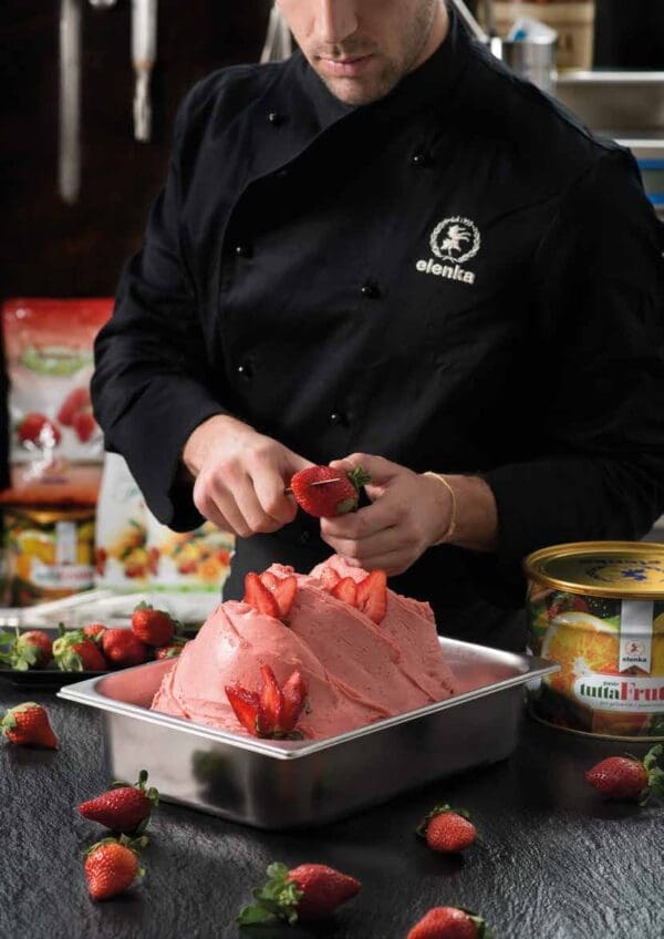 A chef in a black uniform is preparing a strawberry dessert. He is cutting a fresh strawberry over a tray filled with strawberry ice cream, which is decorated with whole strawberries. Various ingredients and a tin can are visible in the background.