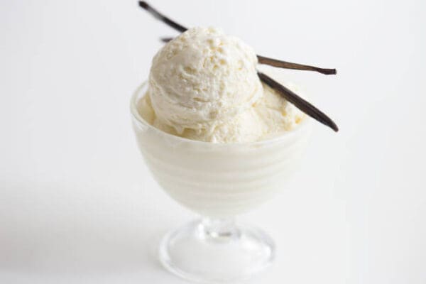 A glass bowl containing two scoops of vanilla ice cream, made silky smooth with Elenka Cremox 298 Ice Cream Emulsifier and Stabilizer (20x1Kg case), garnished with two vanilla bean pods, all set against a white background.