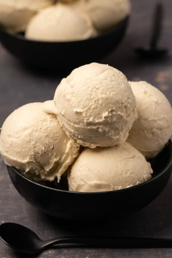 A black bowl brimming with several scoops of creamy vanilla gelato made from Elenka Hipsoya Low Fat Base (Soy Gelato Base) is placed on a dark surface. In the background, another black bowl with more gelato is partially visible. Two black spoons rest nearby, enhancing the minimalistic presentation.