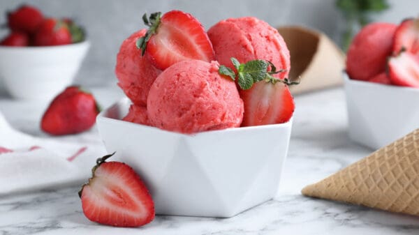 A white geometric bowl filled with scoops of strawberry sorbet, garnished with fresh strawberries and a mint sprig, is set on a marble surface. In the background, there are additional bowls of strawberries and an empty waffle cone.