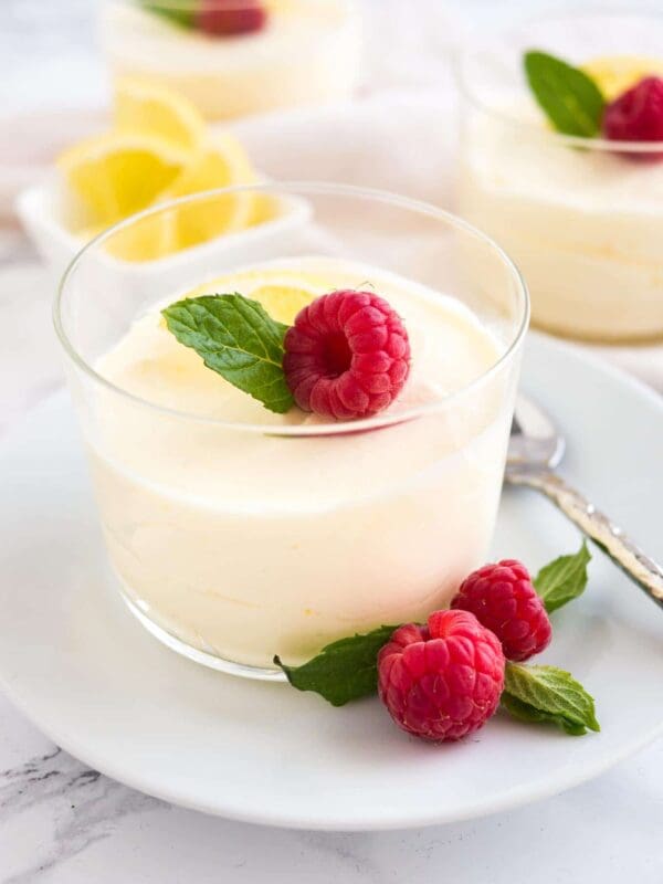 A glass cup of lemon mousse, created using Elenka Base Montelenka Vegetable Cream from a 20x1Kg case, is topped with a raspberry and mint leaf. It sits gracefully on a white plate adorned with additional raspberries and mint leaves. A spoon lies nearby, with another serving and slices of lemon visible in the background.