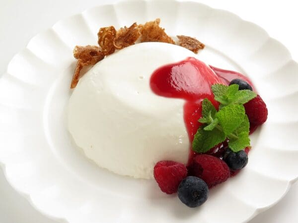 A dessert made with Elenka Base Montelenka Vegetable Cream (20x1Kg case) is elegantly plated, drizzled with red berry sauce and adorned with fresh raspberries, blueberries, and mint leaves. Delicate crumbles of caramelized sugar are thoughtfully arranged behind the dessert for a finishing touch.