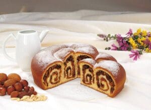 A round, sliced Gubana cake is dusted with powdered sugar, revealing a spiral filling. It's placed on a white surface beside a small white pitcher, a mix of nuts, and colorful wildflowers.