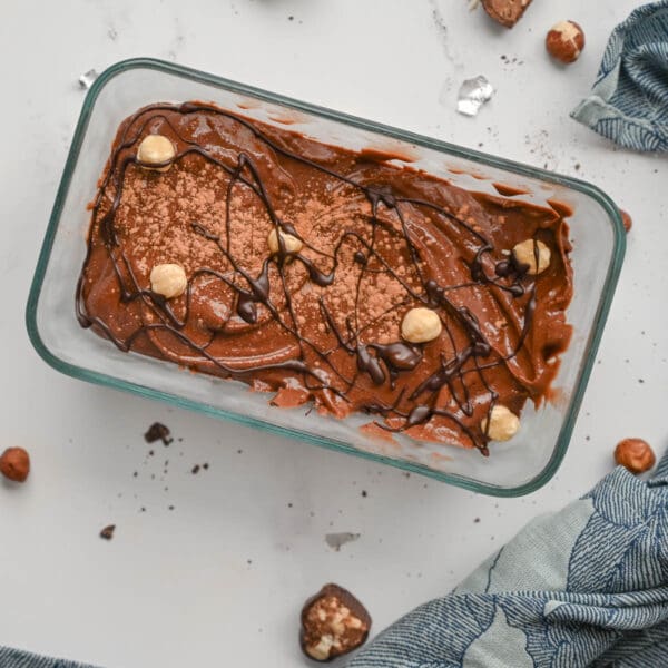 A glass dish filled with rich chocolate mousse, topped with hazelnuts and Elenka Baci Italian Kiss Hazelnut-Nut Pastes #430, garnished with cocoa powder and drizzled chocolate. Surrounded by whole hazelnuts, nut pastes, tiny chocolate shards, and a blue-patterned cloth.