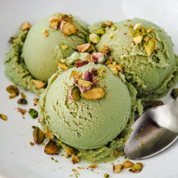 Three scoops of Elenka Pistachio P ice cream (4x2.5Kg), topped with chopped pistachios, sit elegantly on a white plate. A silver spoon rests beside them, poised to dive in.