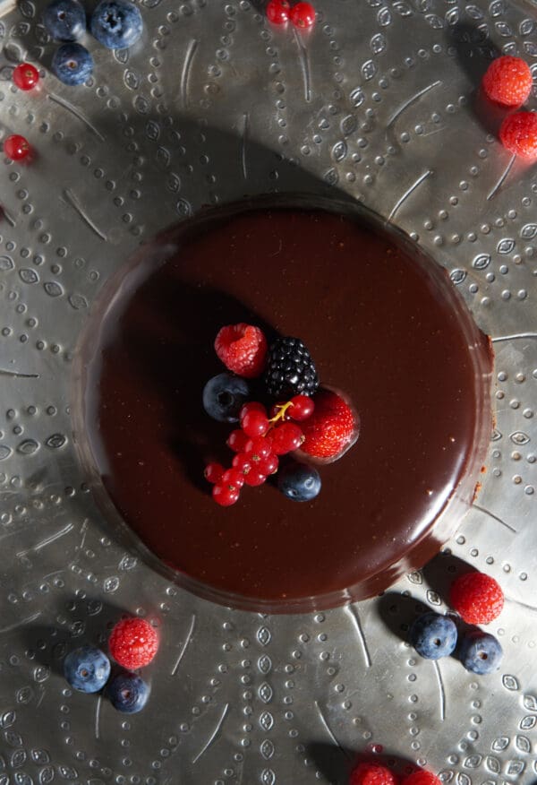 A glossy chocolate cake is topped with assorted raspberries, blueberries, blackberries, and currants, featuring Luxardo Raspberry Jams and Spreads drizzles on a textured metallic surface adorned with scattered berries.